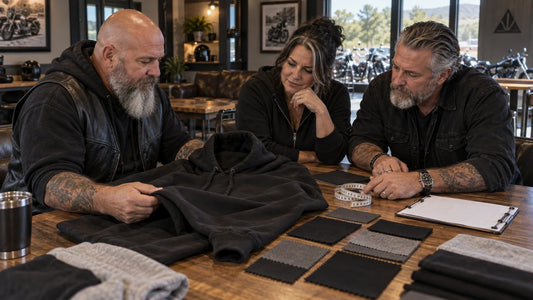 Three mature club organizers review a black hoodie blank, fabric swatches and fit measurements at a sample table.