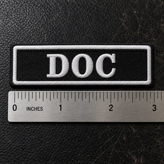 Compact 3 by 1 inch motorcycle name bar beside a precise metal ruler on a leather workbench for clear scale, showing custom motorcycle club name bar patch options.
