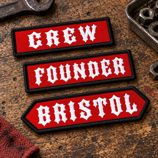 Custom-shaped short club-approved identifier bars arranged on a dark leather inspection surface, fully visible with realistic horizontal proportions, showing custom motorcycle club name bar patch options.