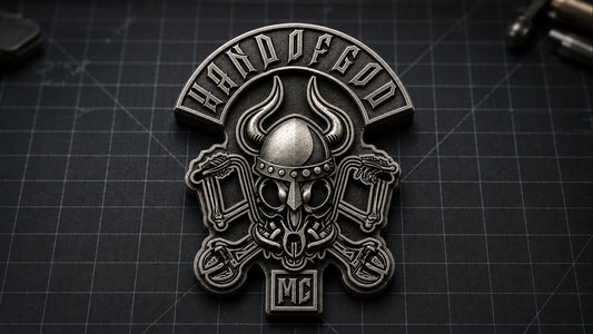Wide workshop view of a custom-outline antique silver HAND OF GOD pin with a sculpted Viking skull and pistons on a drafting mat.