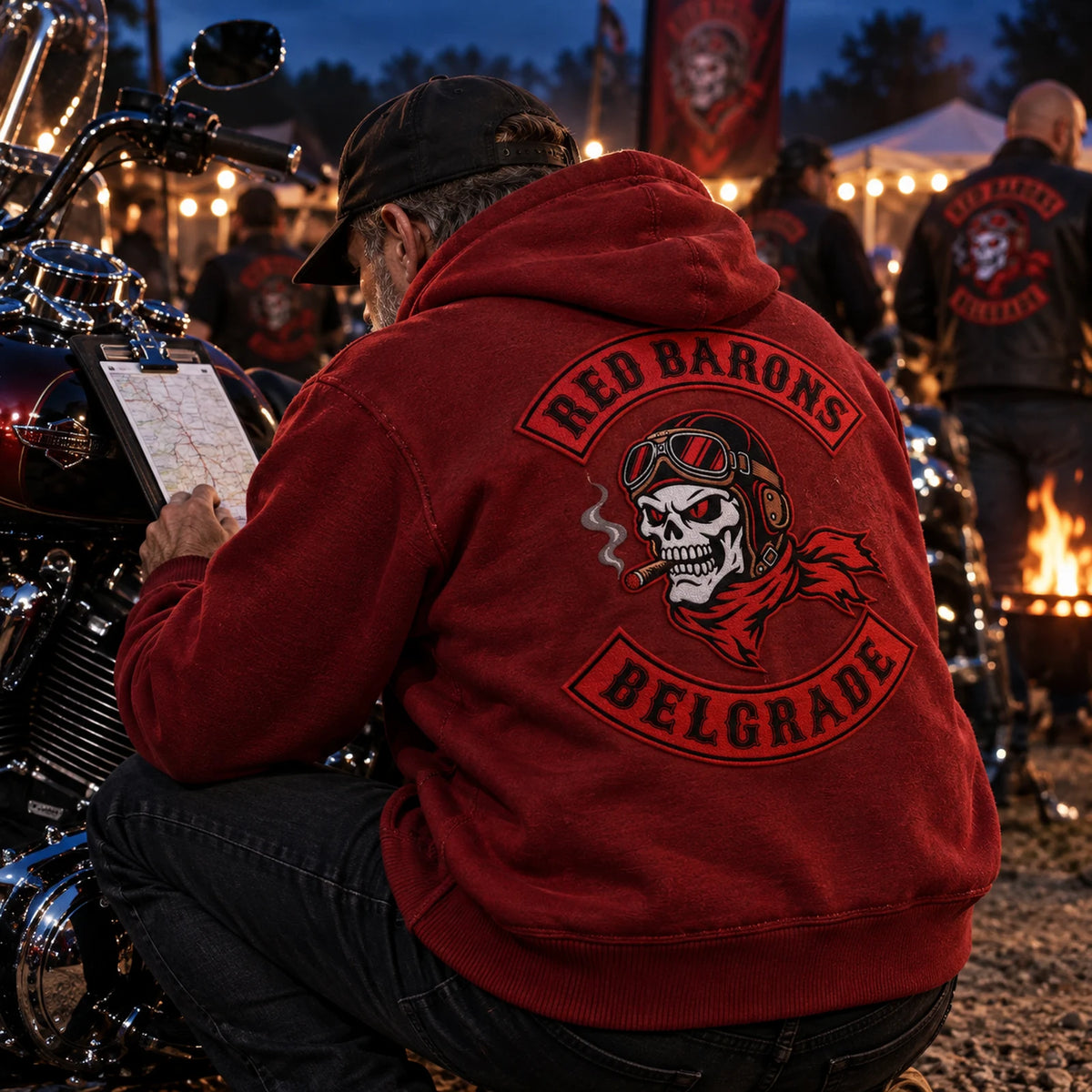 Rider in a deep red motorcycle club hoodie checking a rally route beside a V-twin motorcycle.
