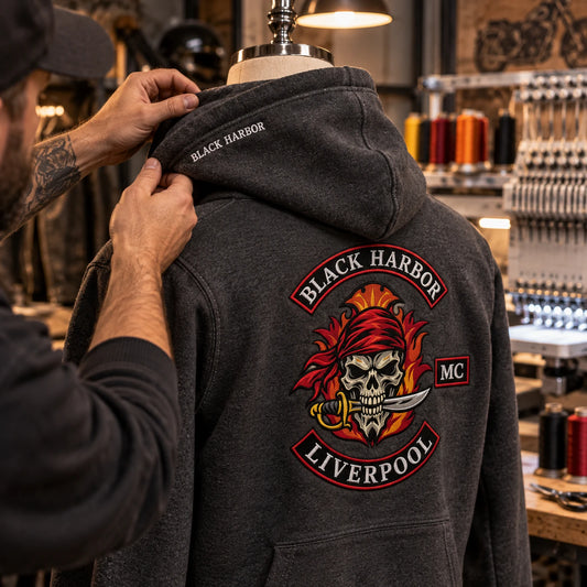 Craftsperson inspecting small Black Harbor embroidery on the hood of a charcoal motorcycle club hoodie.