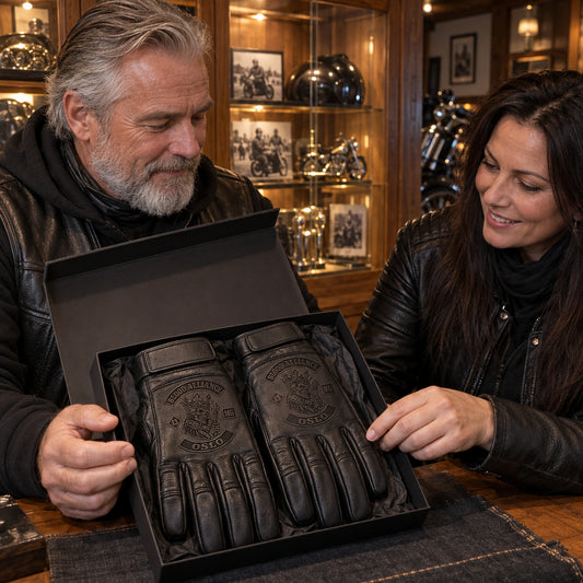 Matte black full-finger cowhide leather glove pair with restrained motorcycle club branding in wood-and-glass clubhouse presentation cabinet.