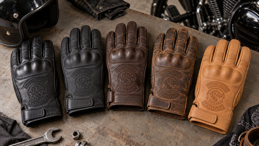 Five complete cowhide leather glove samples in black, matte black, dark brown, vintage brown and natural tan with restrained motorcycle club branding in large warm-grey studio worktable with five separate leather swatches.
