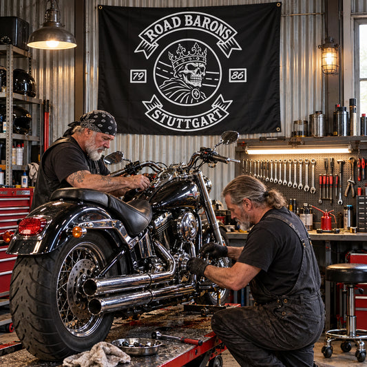 Two mechanics service a V-twin motorcycle beneath a custom Road Barons square workshop flag.