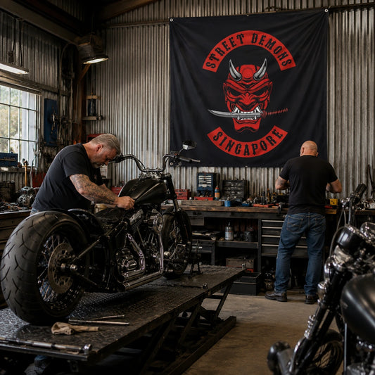 Two mechanics work on a V-twin motorcycle beneath a custom square club flag in a garage.