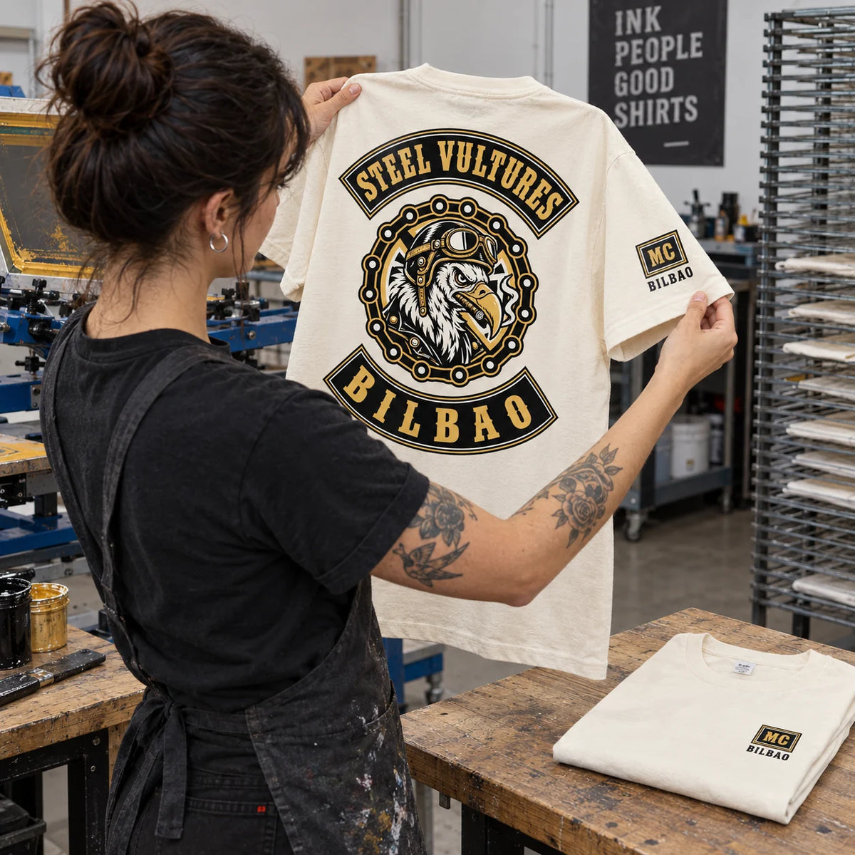 Printmaker inspecting a natural off-white Steel Vultures Bilbao club T-shirt with a small Bilbao MC sleeve mark in a professional print studio.