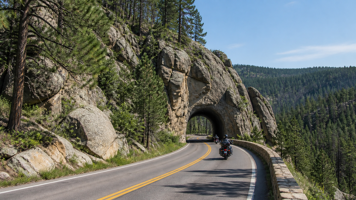 Motorcyclists riding a curving Black Hills road through granite formations and pine forest
