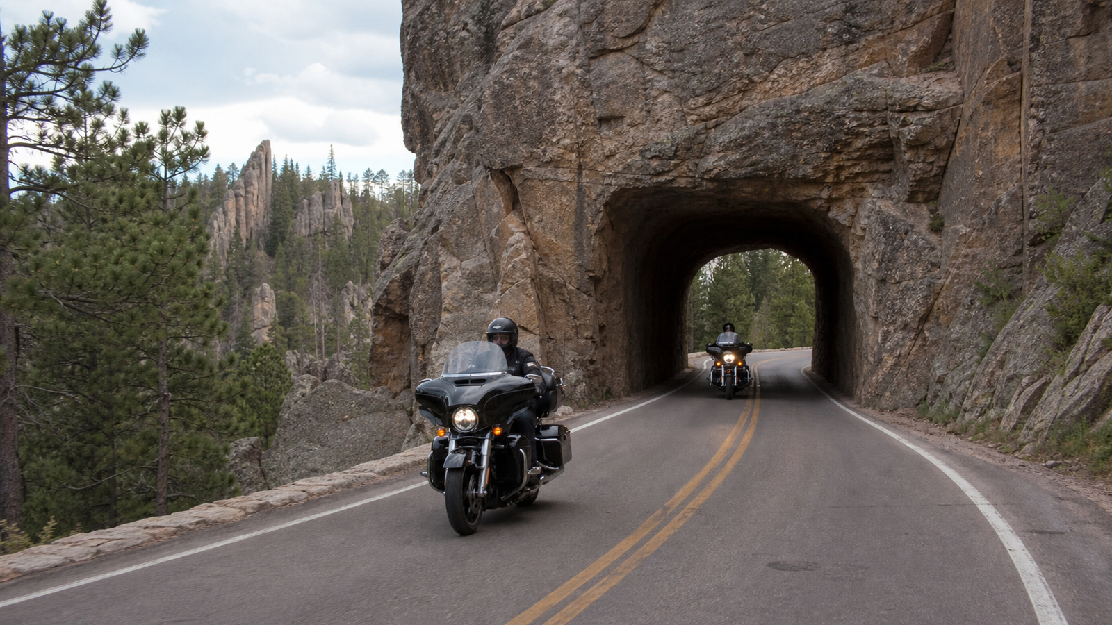 Two touring motorcyclists maintaining safe spacing beside a narrow granite tunnel in the Black Hills