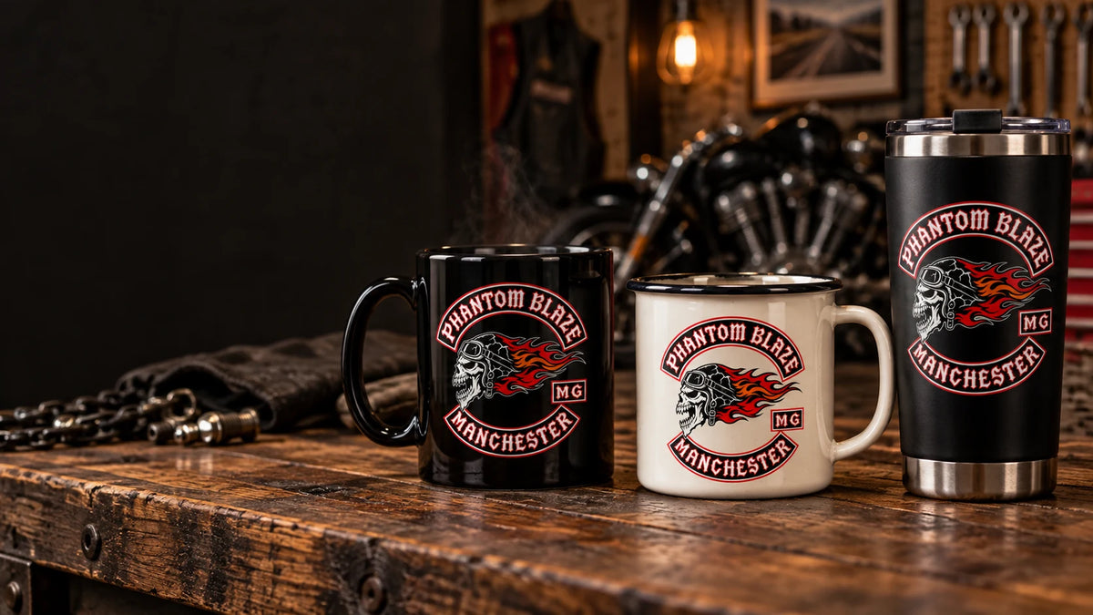 Wide premium clubhouse hero still life with a black ceramic mug, cream enamel camp mug and black stainless travel mug grouped on a heavy timber counter; each product carries the supplied club emblem; subtle unbranded V-twin workshop details in soft focus and generous dark negative space on the left.