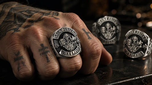 Tattooed biker hand wearing a heavy custom motorcycle club ring on the middle finger, with matching silver rings blurred in the background.