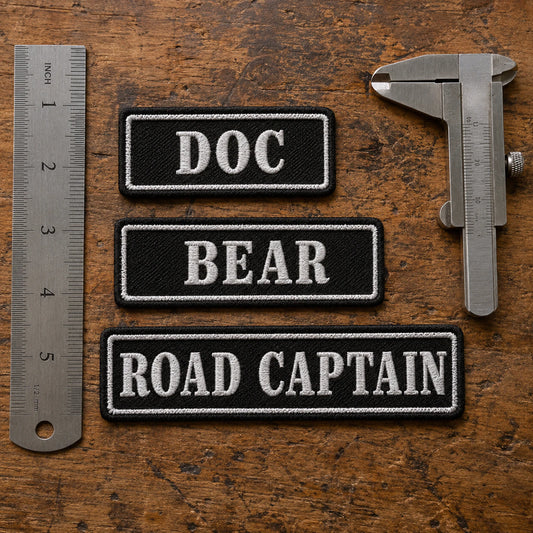 Several true-scale custom-width name bars beside calipers and a ruler on a patch inspection table, with no hands present, showing custom motorcycle club name bar patch options.