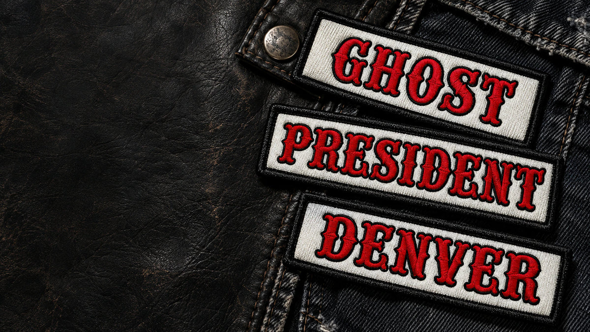 A premium horizontal arrangement of several coordinated road-name, officer-title and chapter name-bar patches on worn black leather and dark denim, with realistic scale, visible embroidery details and open space on the left for website text, showing custom motorcycle club name bar patch options.