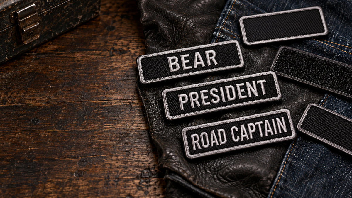 A premium horizontal arrangement of several coordinated road-name, officer-title and chapter name-bar patches on worn black leather and dark denim, with realistic scale, visible embroidery details and open space on the left for website text, showing custom motorcycle club name bar patch options.