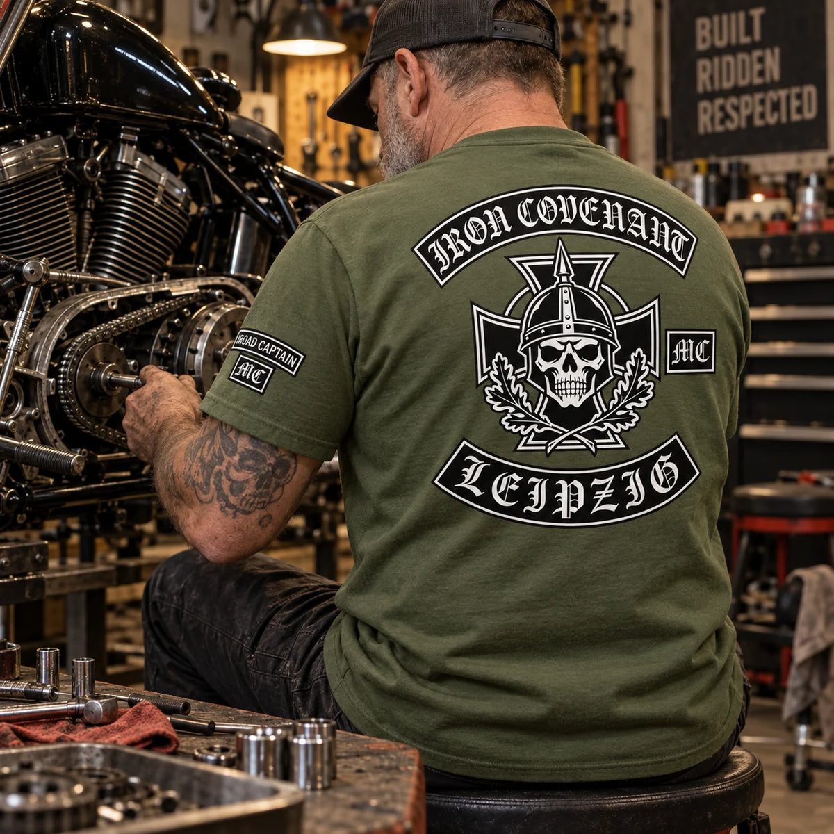 Iron Covenant Leipzig mechanic wearing a military-green club T-shirt with a small Road Captain sleeve identifier while servicing a V-twin motorcycle.