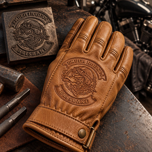 Tan cowhide full-finger leather glove with a smooth stable decoration panel with restrained motorcycle club branding in blackened steel leatherworking bench with a plain embossing die nearby.