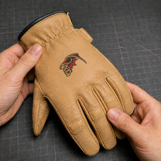 A tan motorcycle club glove carries a compact full-color printed emblem between the knuckles and wrist gathering.