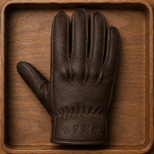 A brown leather motorcycle club glove shows the supplied HPMC initials pressed directly into its fixed cuff without a separate patch.