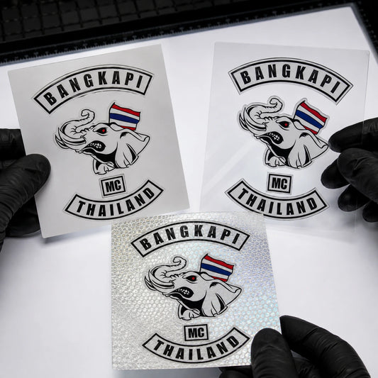 Production specialist comparing vinyl, transparent and reflective motorcycle club sticker material swatches under inspection lights.