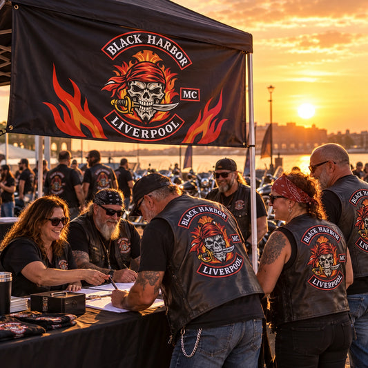 Mature V-twin riders gather at a rally tent displaying a custom horizontal motorcycle club flag.