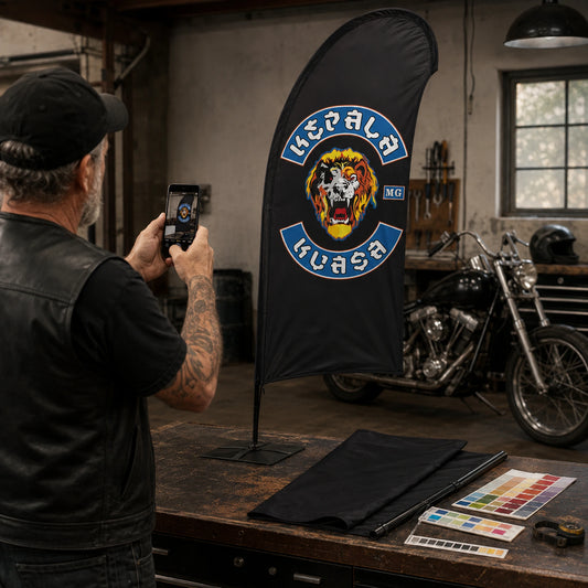 Send Your Club Artwork: custom motorcycle club feather flag shown in mature rider photographing an authorized club emblem on a clean garage workbench.