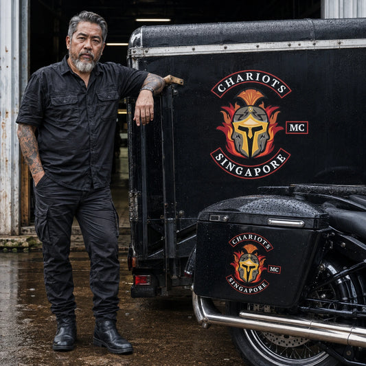 Road captain and mechanic beside a rain-damp support trailer and pannier carrying durable custom vinyl club stickers.