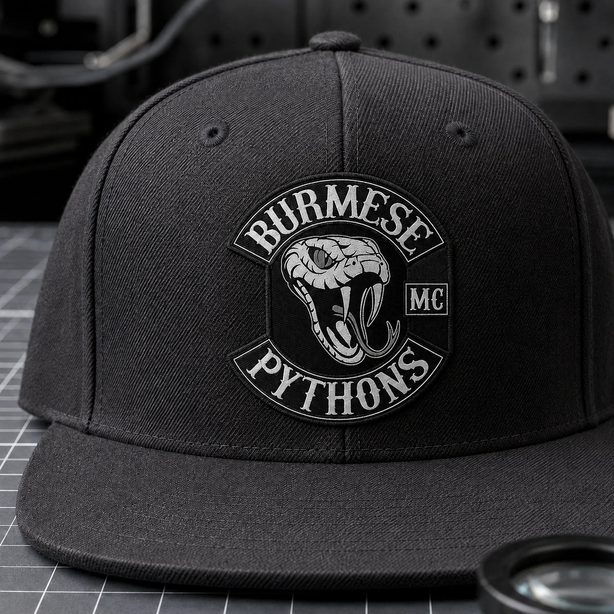 A charcoal motorcycle club flat-brim cap displays a finely detailed woven emblem patch.